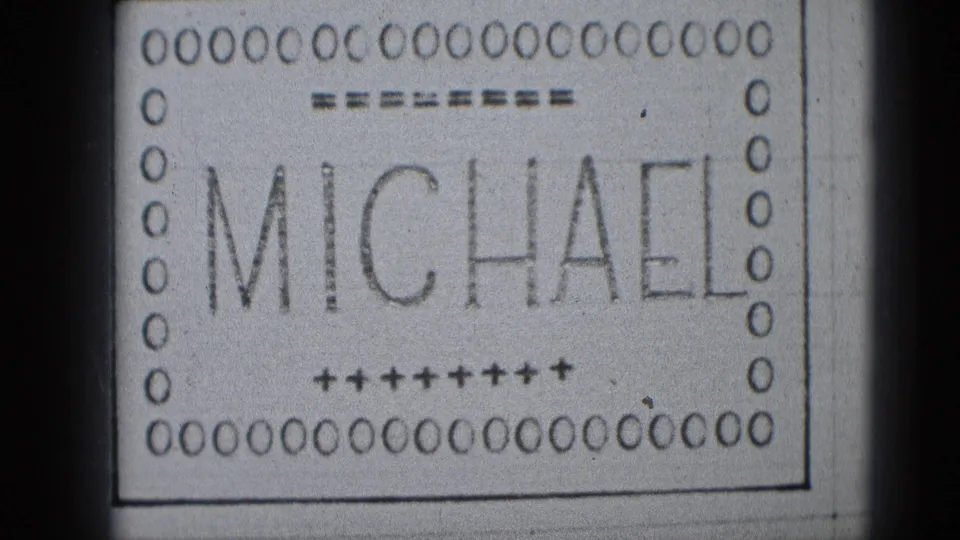Close 8mm frame of the MICHAEL title card, with the lower border and plus-sign decoration visible in black and white.