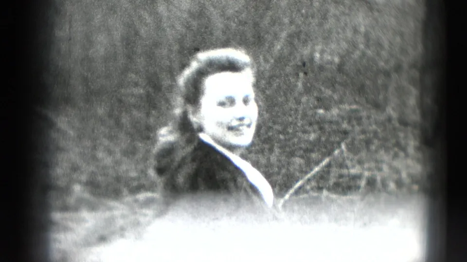 The woman smiles toward the camera in a grainy 8mm frame, with her dark coat and outdoor setting visible despite bright flare.