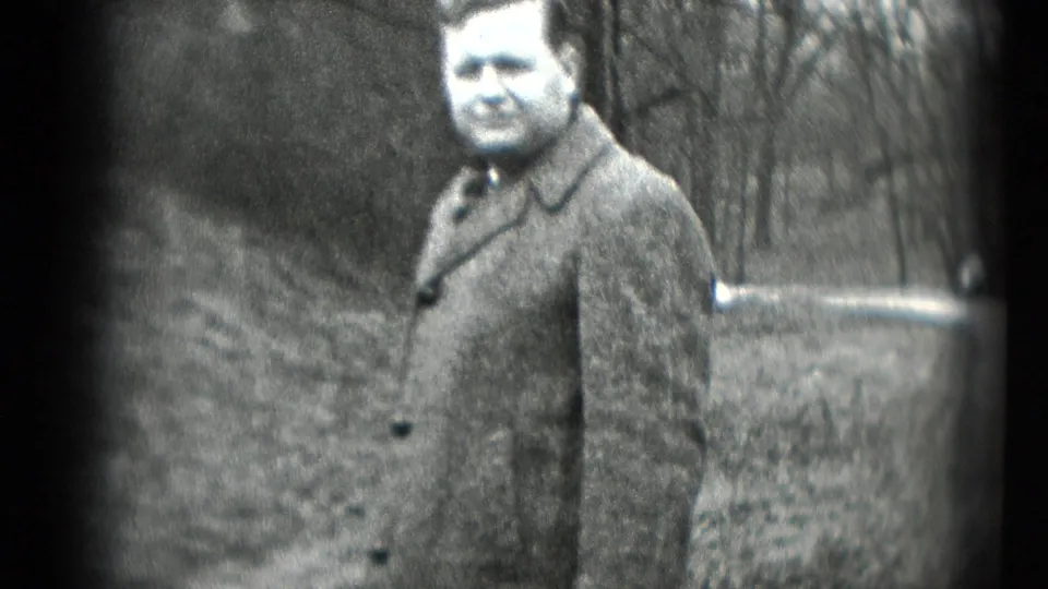 An adult man in a heavy coat stands outdoors near bare trees in grainy black-and-white home-movie footage from Middletown, Pennsylvania.