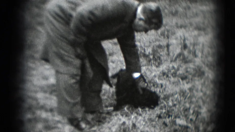 A man in a long coat reaches down toward a small dark dog in a wooded Middletown setting, with soft 8mm focus and dark frame edges.
