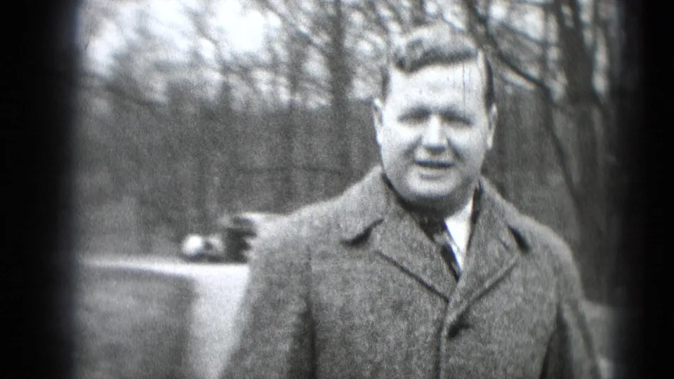 Man in a wool coat faces the camera outdoors, with bare trees and a partial car behind him in 1948 Middletown footage.