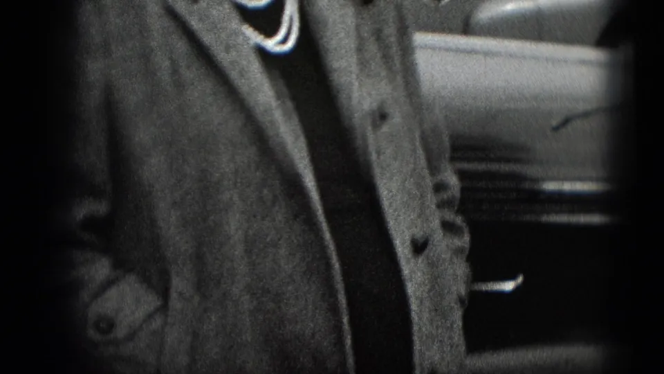 Tight home-movie frame showing a woman's coat and necklace with part of a parked vehicle at the edge.