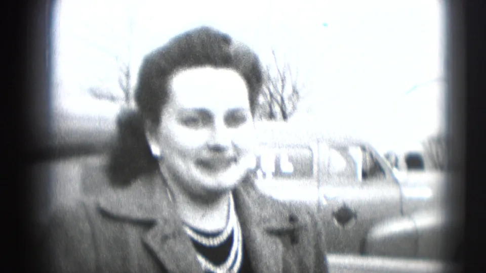 Brightly exposed 8mm frame of the woman smiling, with the street and parked car still visible behind her.