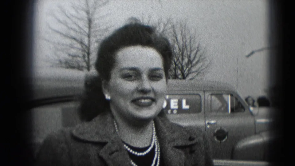 Smiling woman in black-and-white 8mm footage, framed by a parked car, bare trees, and soft vignetted edges.