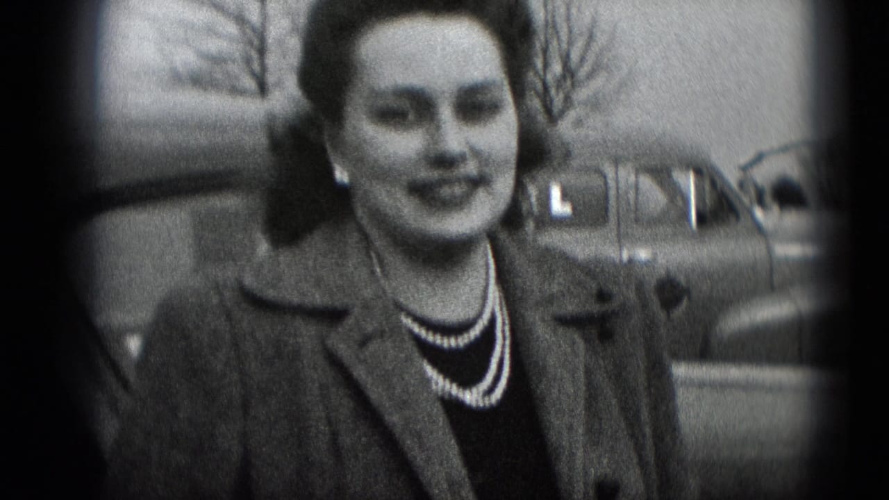 The camera holds on the woman's face, coat, and pearl necklace, with a rounded car and leafless branches visible in the background.