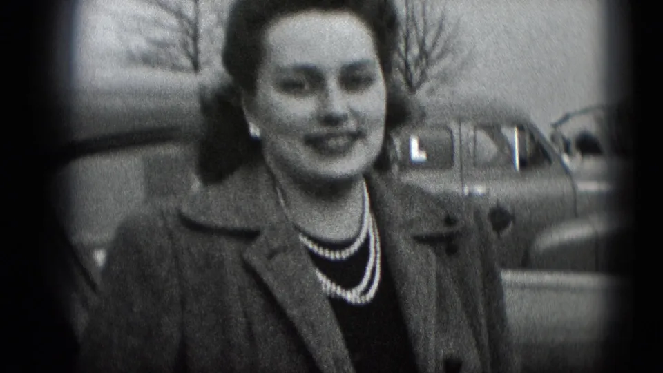 The camera holds on the woman's face, coat, and pearl necklace, with a rounded car and leafless branches visible in the background.