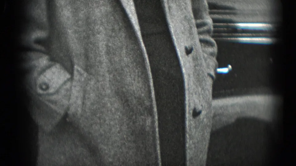 The camera drops to the woman's coat, dark dress, and hands near her pockets, with part of a parked automobile behind her.