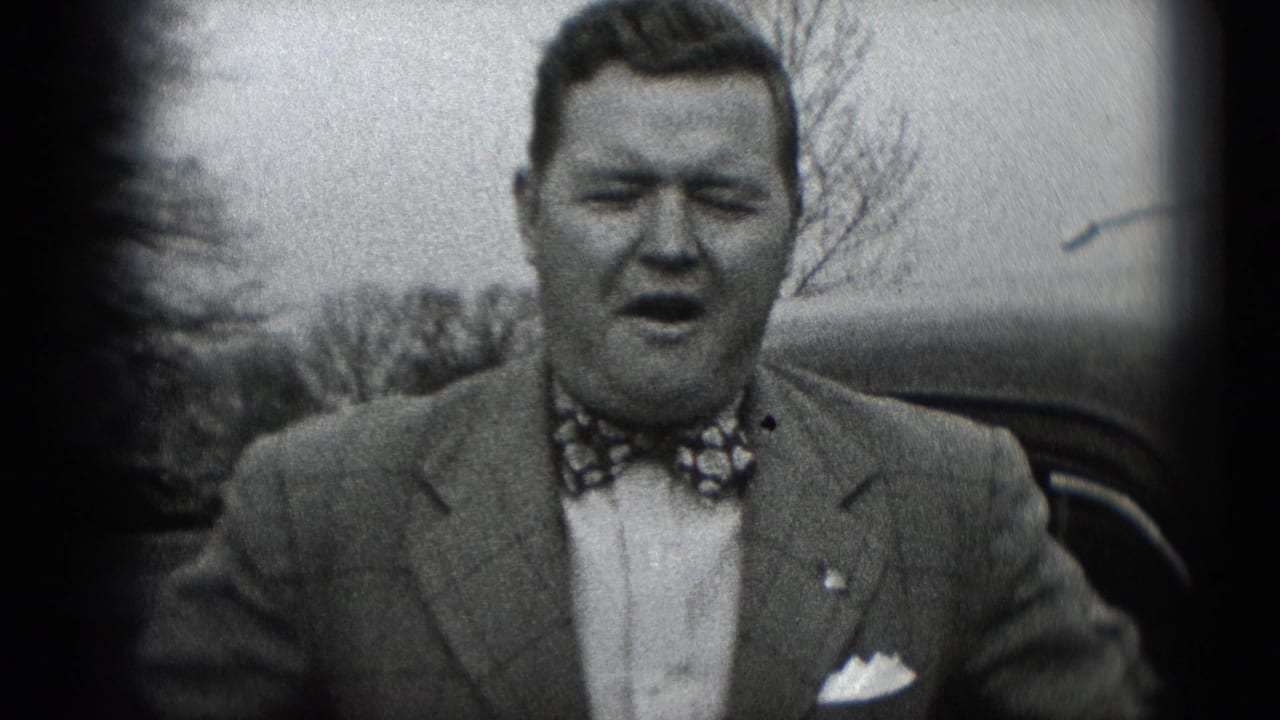 A man in a suit, patterned bow tie, and pocket square faces the camera outdoors, with trees and a dark automobile behind him.
