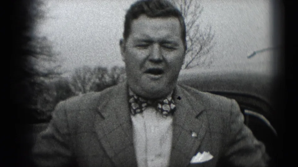 A man in a suit, patterned bow tie, and pocket square faces the camera outdoors, with trees and a dark automobile behind him.