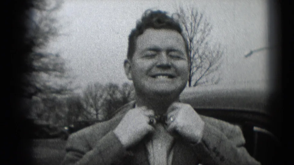 A smiling adult man in a suit adjusts his patterned bow tie in a black-and-white 8mm home-movie frame from Middletown, Pennsylvania.
