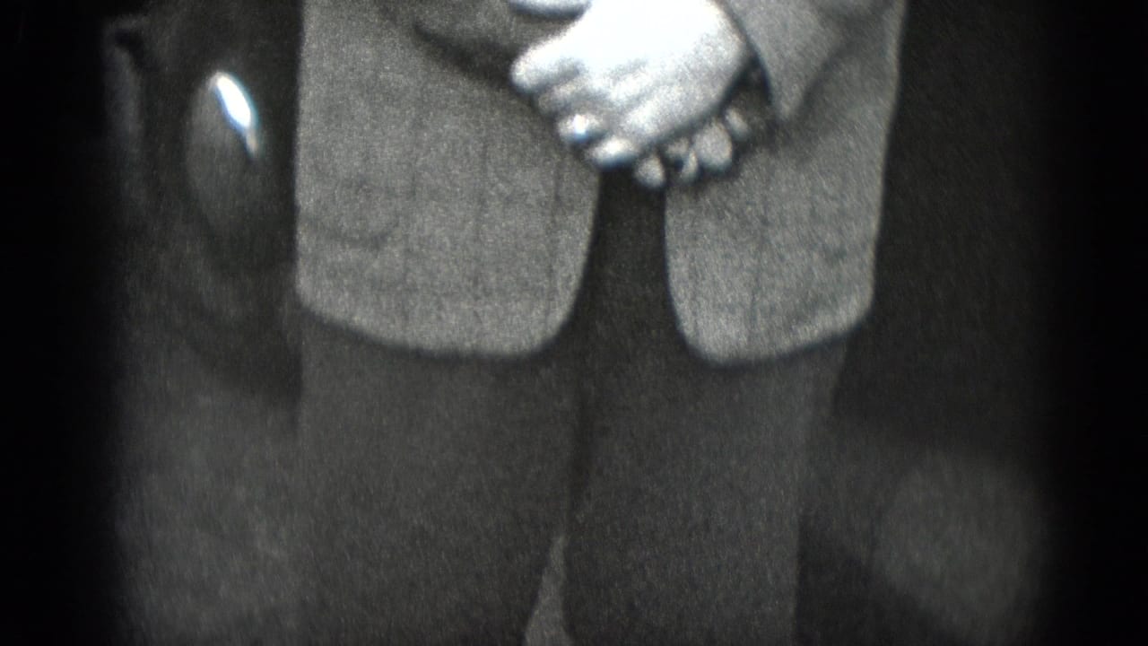 Close view of a suited man's clasped hands and lap beside a dark car interior.