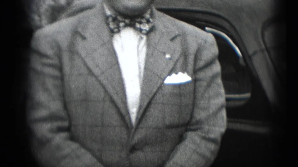A suited adult man with patterned bow tie and pocket square sits beside a car in black-and-white 8mm home-movie footage.