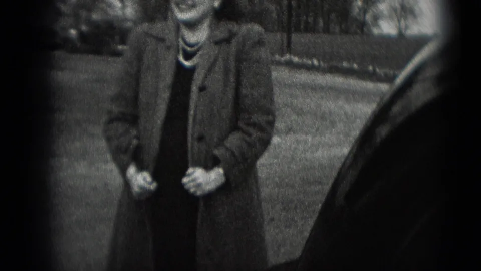 A woman in a long coat and pearl necklace stands outdoors beside a car in 1948 Middletown home-movie footage.