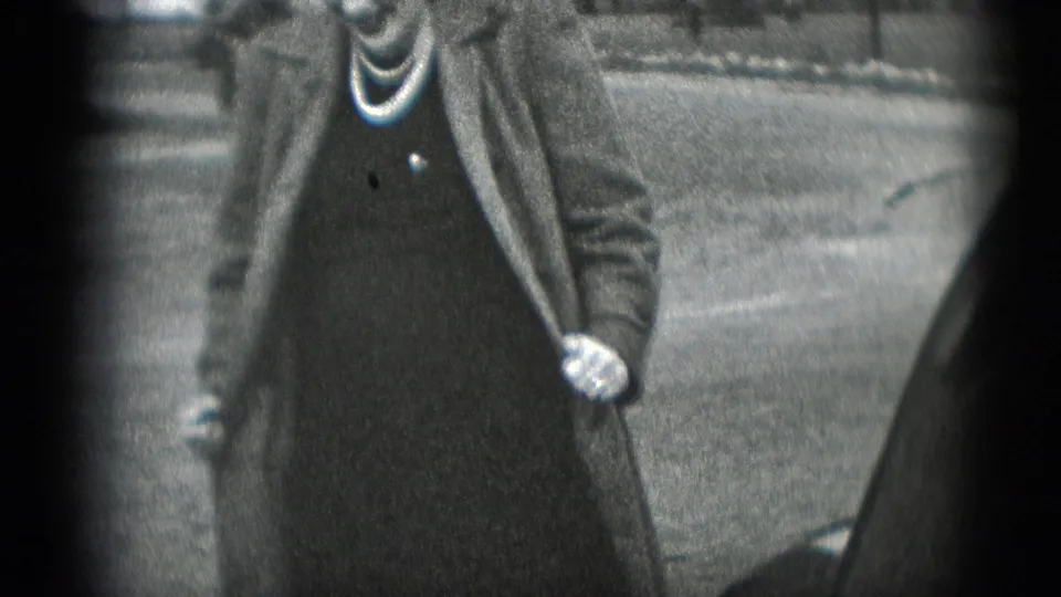 The woman opens her long coat to show a dark dress while standing outdoors in the 1948 Middletown home movie.