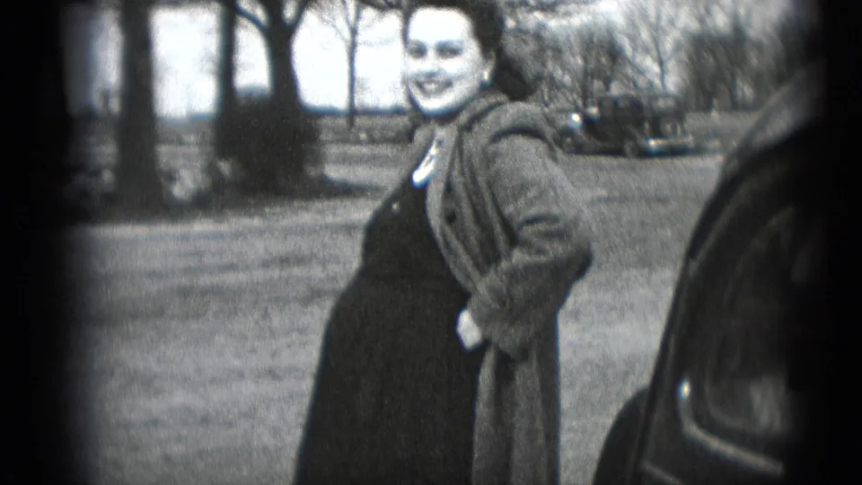 The woman remains posed beside the automobile, smiling in a soft 8mm frame with trees and lawn visible.