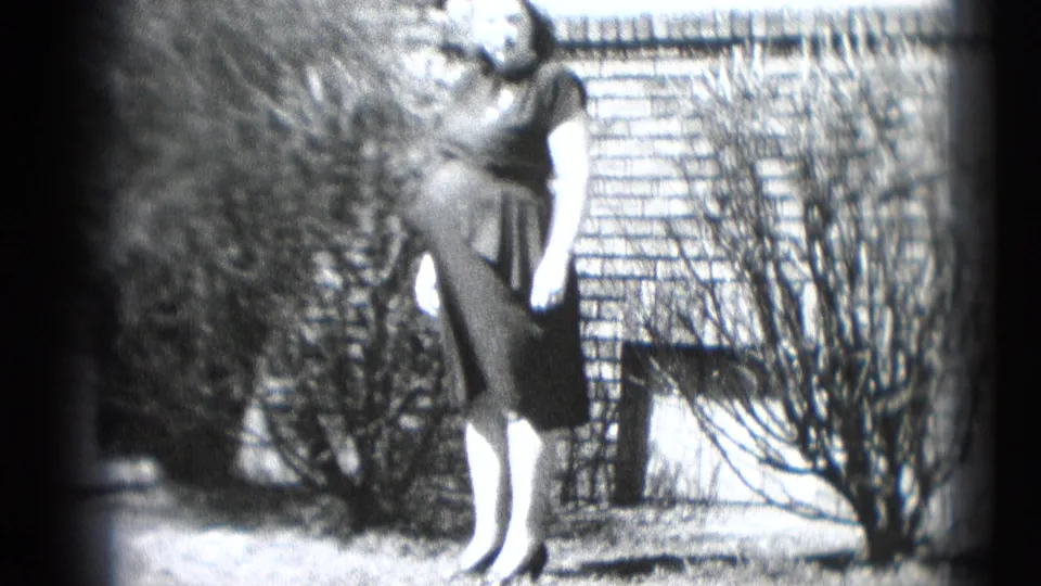 The same woman appears near a brick wall and shrubs, standing in a dark dress in the Middletown home-movie scene.
