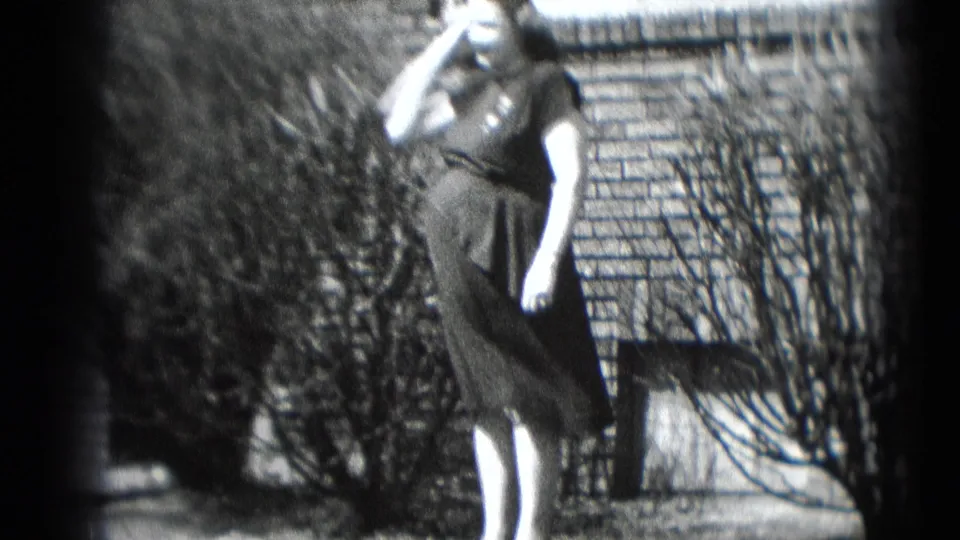 The 1948 home-movie frame shows the woman from the side in a yard, holding the receiver as she stands near shrubs and brickwork.