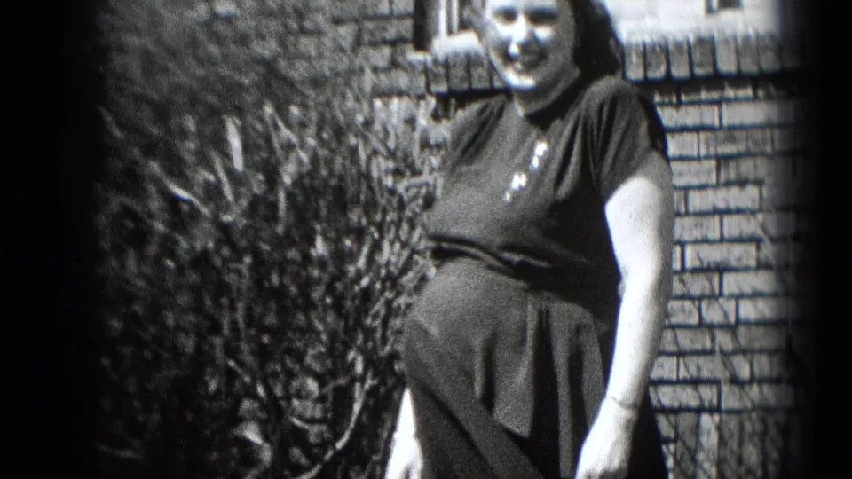 In Middletown, Pennsylvania home-movie footage, a pregnant woman in a dark dress smiles outdoors near a brick wall and shrubs.