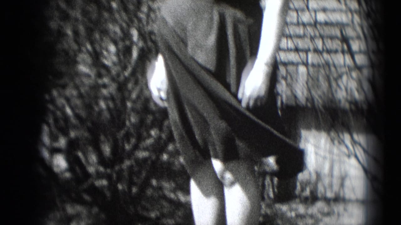 The woman stands near shrubs and brickwork as wind spreads the fabric of her dark dress in the 8mm footage.