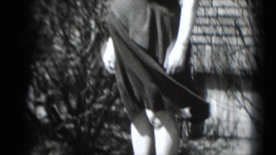 The woman stands near shrubs and brickwork as wind spreads the fabric of her dark dress in the 8mm footage.