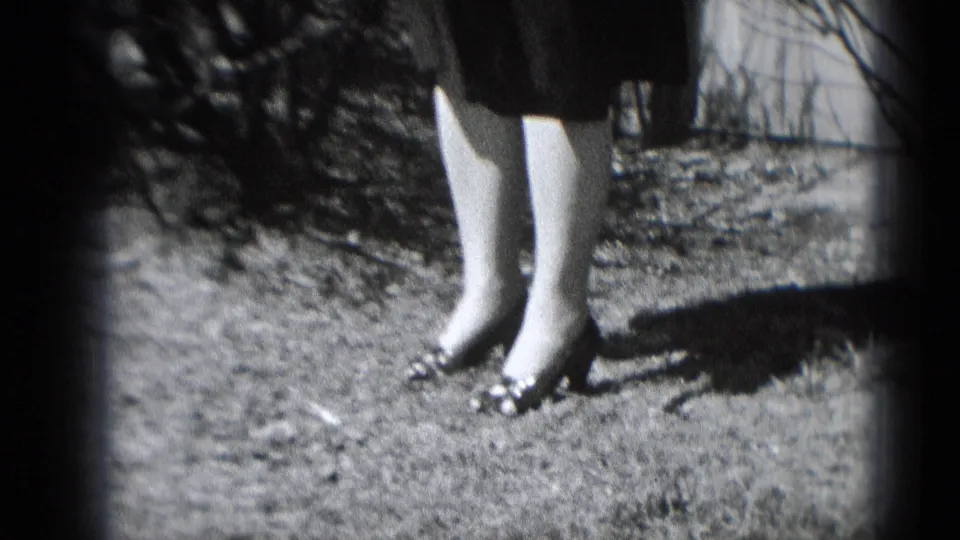 The camera frames the woman's dress hem, legs, and heeled shoes on sunlit ground near shrubs and a brick exterior.