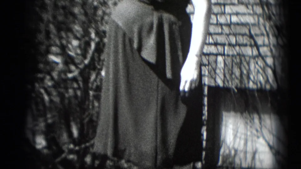 A closer home-movie view shows the woman's dark dress, arm, brick wall, and nearby bare branches in strong sunlight.