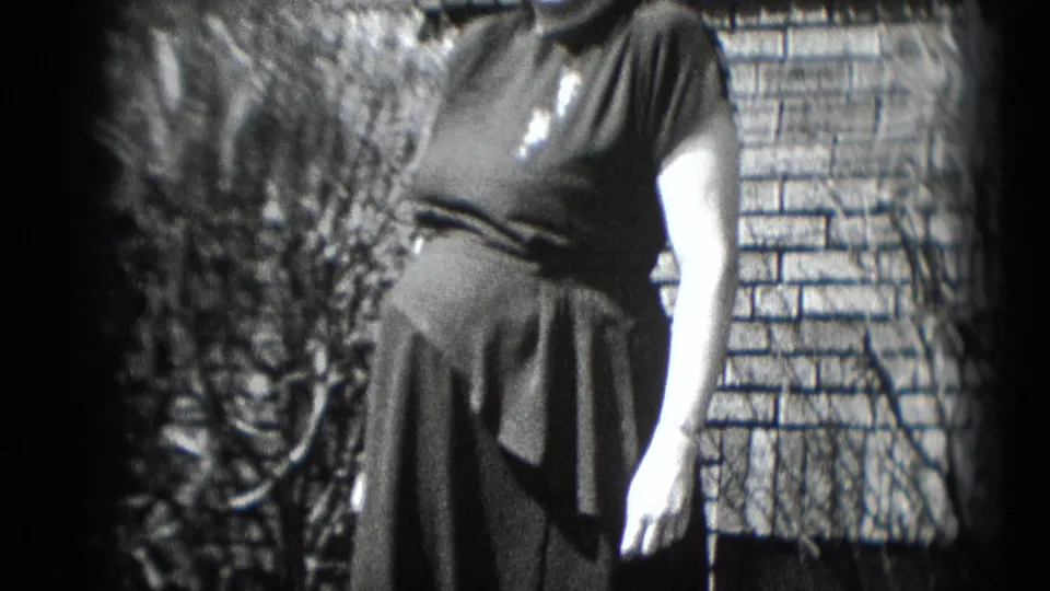 Pregnant woman in a dark dress poses outside a brick house in metadata-identified Middletown, Pennsylvania archival home-movie footage.