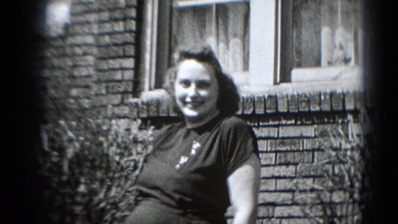 Smiling pregnant woman faces the camera near a brick wall and window in black-and-white 8mm home-movie footage.