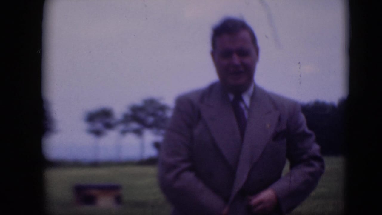 A man in a gray suit faces the camera and adjusts his jacket on grass, with trees and cloudy sky in the background.