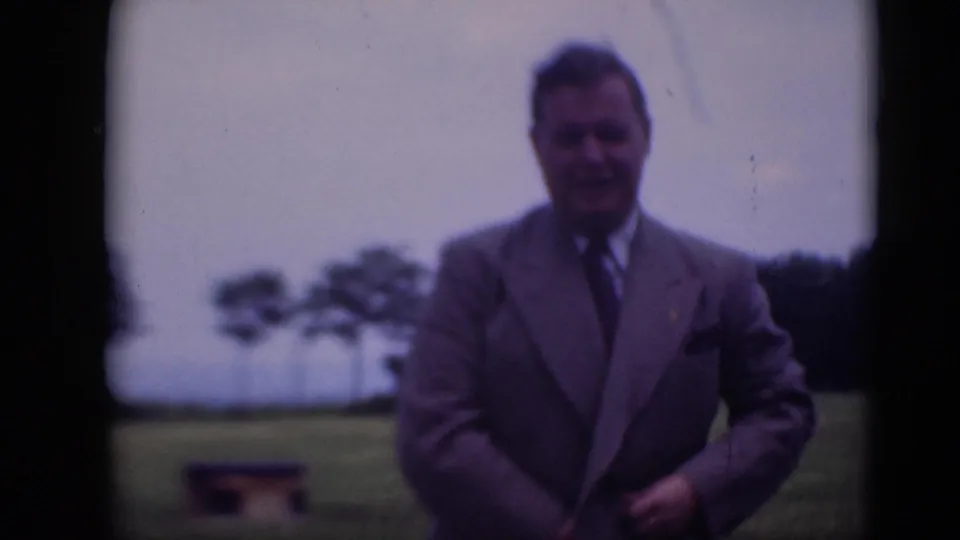A man in a gray suit faces the camera and adjusts his jacket on grass, with trees and cloudy sky in the background.