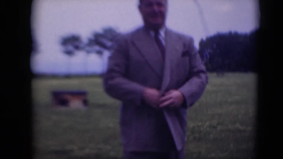 The suited man continues adjusting his jacket in a soft-focus 8mm frame, standing before grass and distant trees.