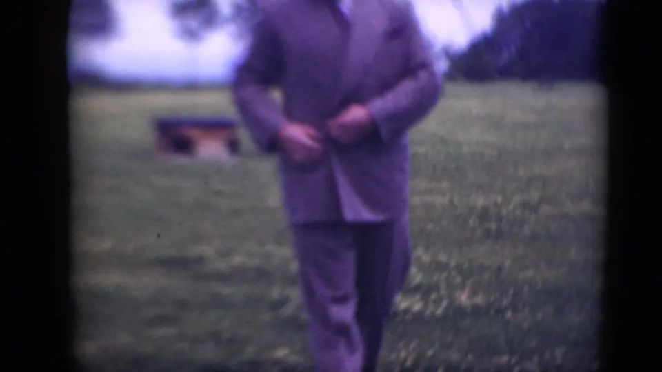 The camera frames the man's suit and legs as he adjusts the jacket, with the grassy field and tree line still visible.