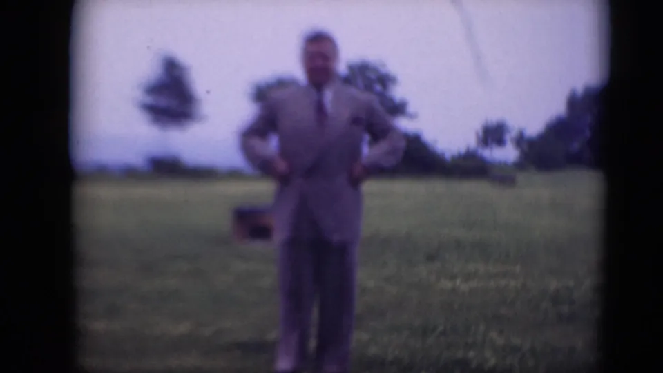 Adult man in a gray suit and tie poses with hands on hips in a grassy field, with trees in the background.