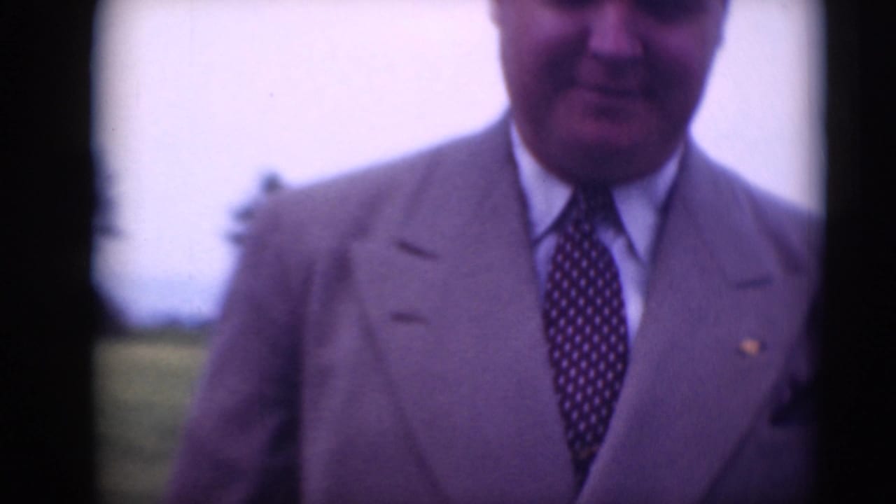 A man in a light suit and patterned tie nears the camera outdoors, with grass and sky behind him.