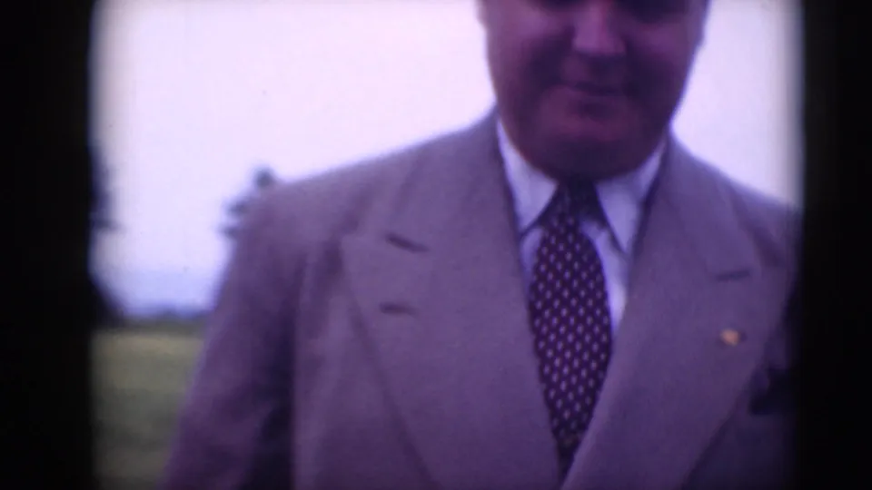 A man in a light suit and patterned tie nears the camera outdoors, with grass and sky behind him.