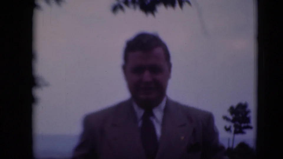 The man in a light suit stands at a distance outdoors, with trees and open sky in the background.