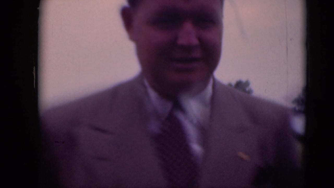 Closer soft-focus view of a man in a light suit standing near the camera outdoors.