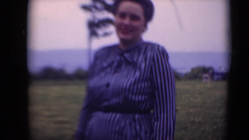 The woman remains framed in the field, smiling at the camera with a tree, hills, and pale sky behind her.