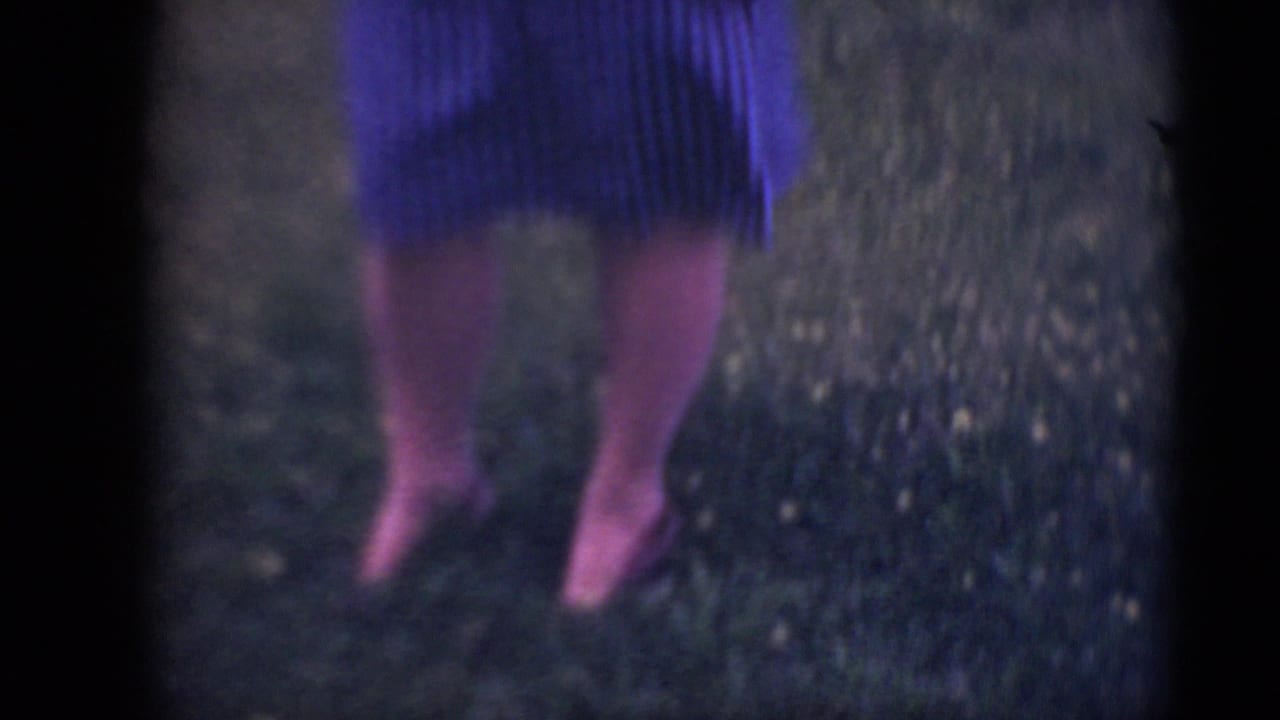 The camera frames the woman's legs and hem of a striped dress as she stands on grass in soft 8mm color.