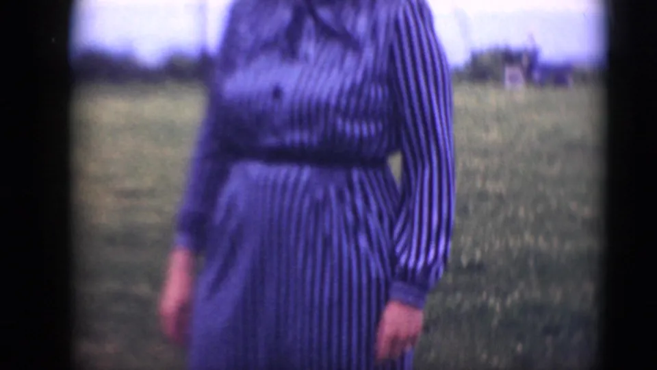 A closer home-movie view shows the woman's belted striped dress and arm against an open grassy background.