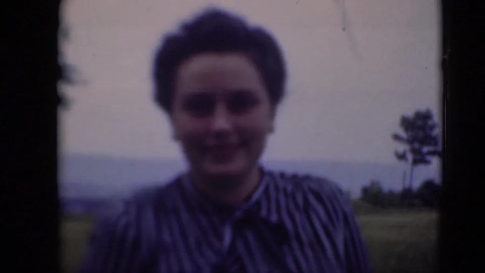 Softly focused 8mm frame of a woman smiling toward the camera on a grassy Middletown lawn, with a tree and horizon in the background.