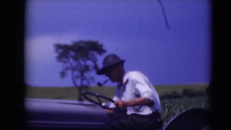 A closer 8mm frame shows the man in profile at the tractor controls, with field and distant trees behind him.
