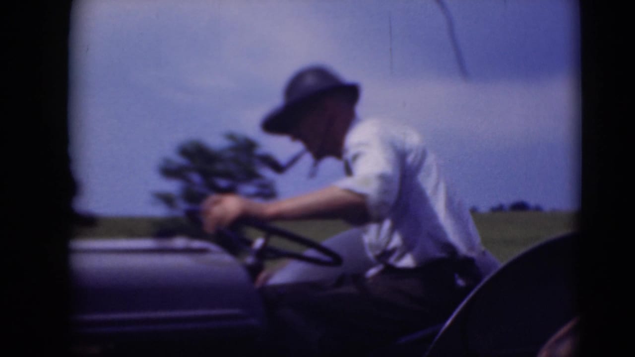 A man in a white shirt and brimmed hat smokes a pipe while steering an open tractor across a rural Pennsylvania field.