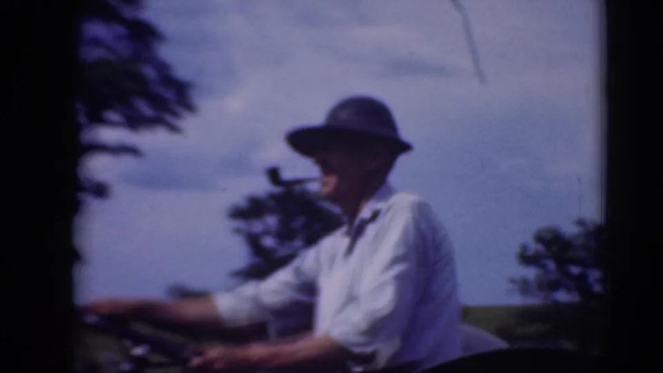 Side view of the tractor driver in 1948 home-movie footage, with his hat, white shirt, pipe, steering wheel, field, and trees visible.