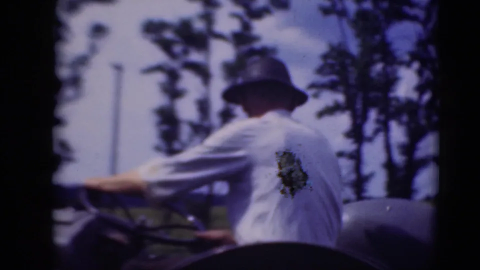 Rear-side view of the man driving the tractor, with his white shirt, hat, steering wheel, nearby trees, and open rural setting visible.