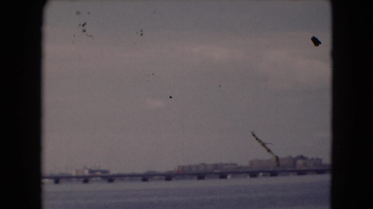 Long bridge crossing a river with a crane-like shape and buildings on the far shore.