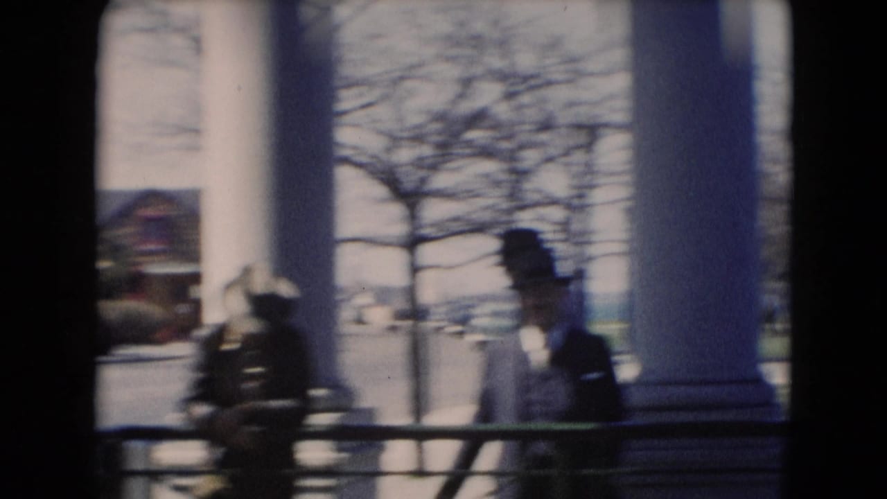 Men in overcoats and hats stand by large white columns near a railing, with trees and houses visible beyond.