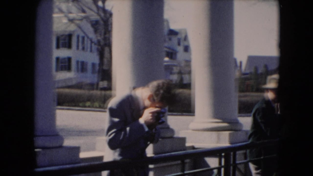 A man appears to adjust a handheld camera by large white columns, with another coated figure at the edge of the frame.
