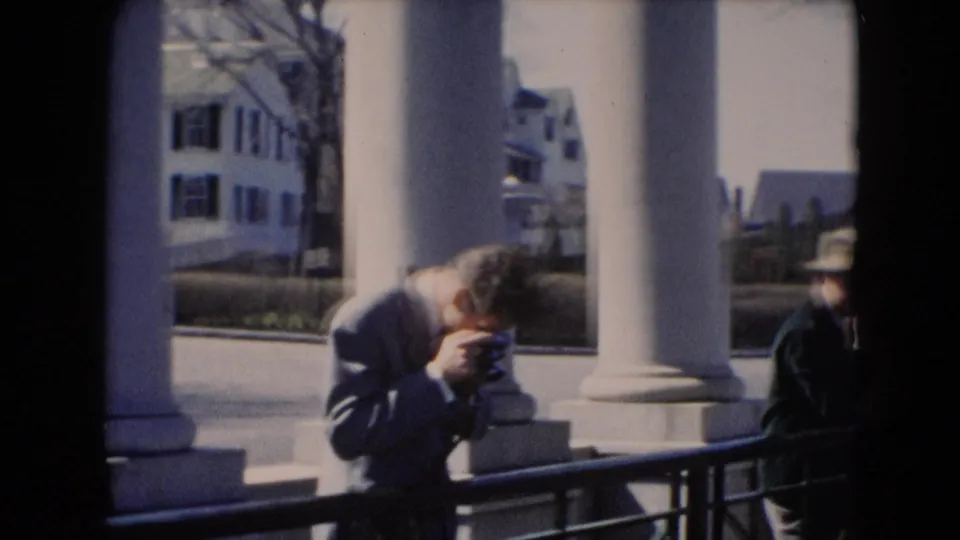 A man appears to adjust a handheld camera by large white columns, with another coated figure at the edge of the frame.