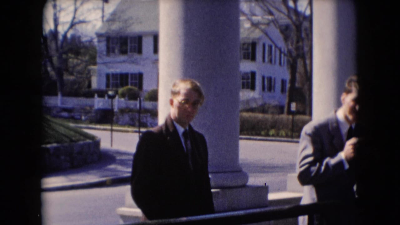 A suited man stands near white porch columns, with another man partly cut off at the edge of the frame.